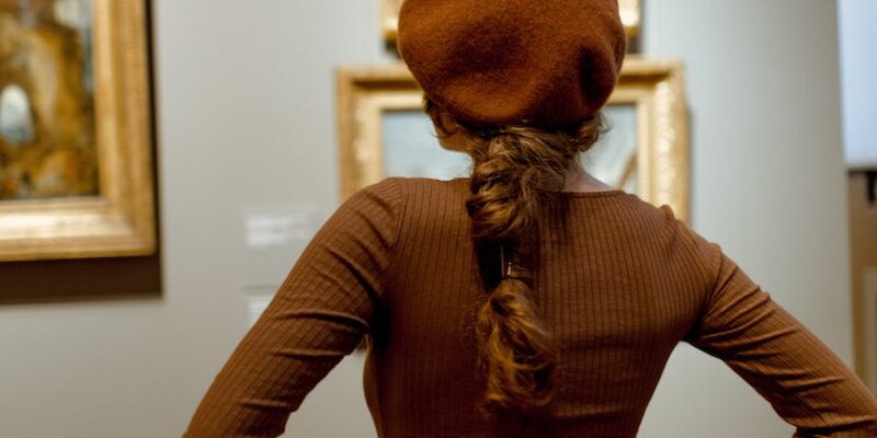 woman in brown long sleeves and hat in front of oil paintings